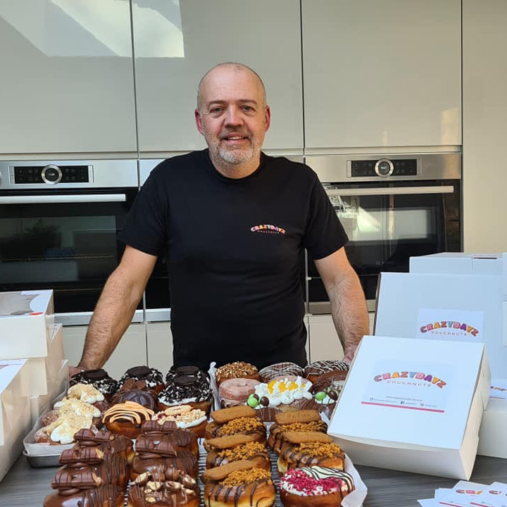 Business owner Neil standing in a kitchen with a range of doughnuts with various toppings