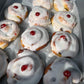 A tray of freshly baked belgian bunstopped with thick white icing and a small red cherry on top, arranged on parchment paper.