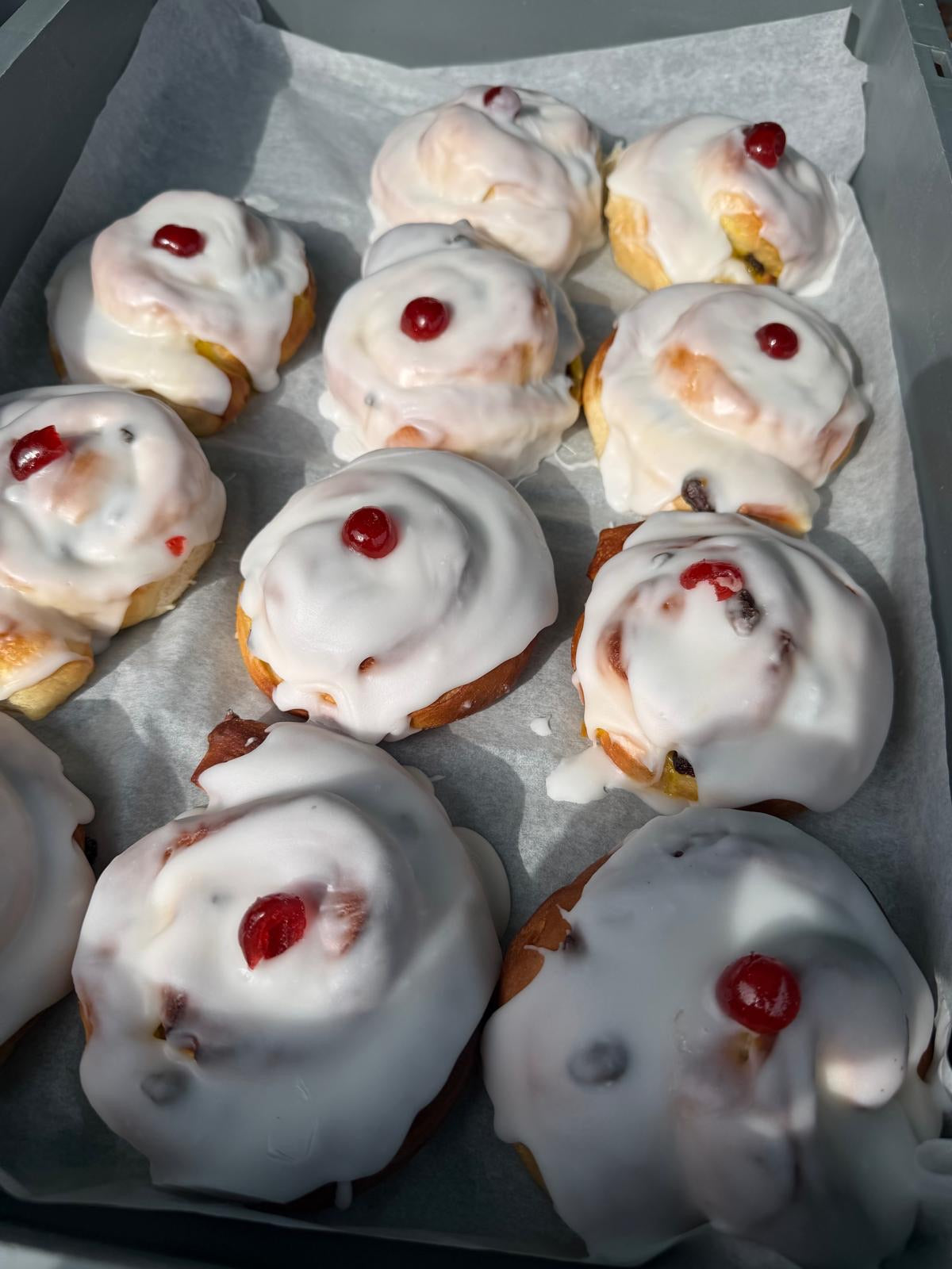 A tray of freshly baked belgian bunstopped with thick white icing and a small red cherry on top, arranged on parchment paper.