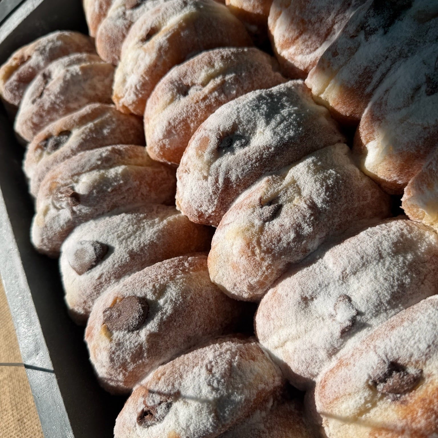 Rows of freshly baked doughnuts, dusted with powdered sugar, arranged tightly in a metal tray.
