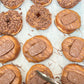 A close-up overhead view of assorted chocolate-topped donuts on a light surface. Several donuts are covered in chocolate sprinkles, while others have glossy chocolate drizzle and a molded chocolate cookie decoration on top.