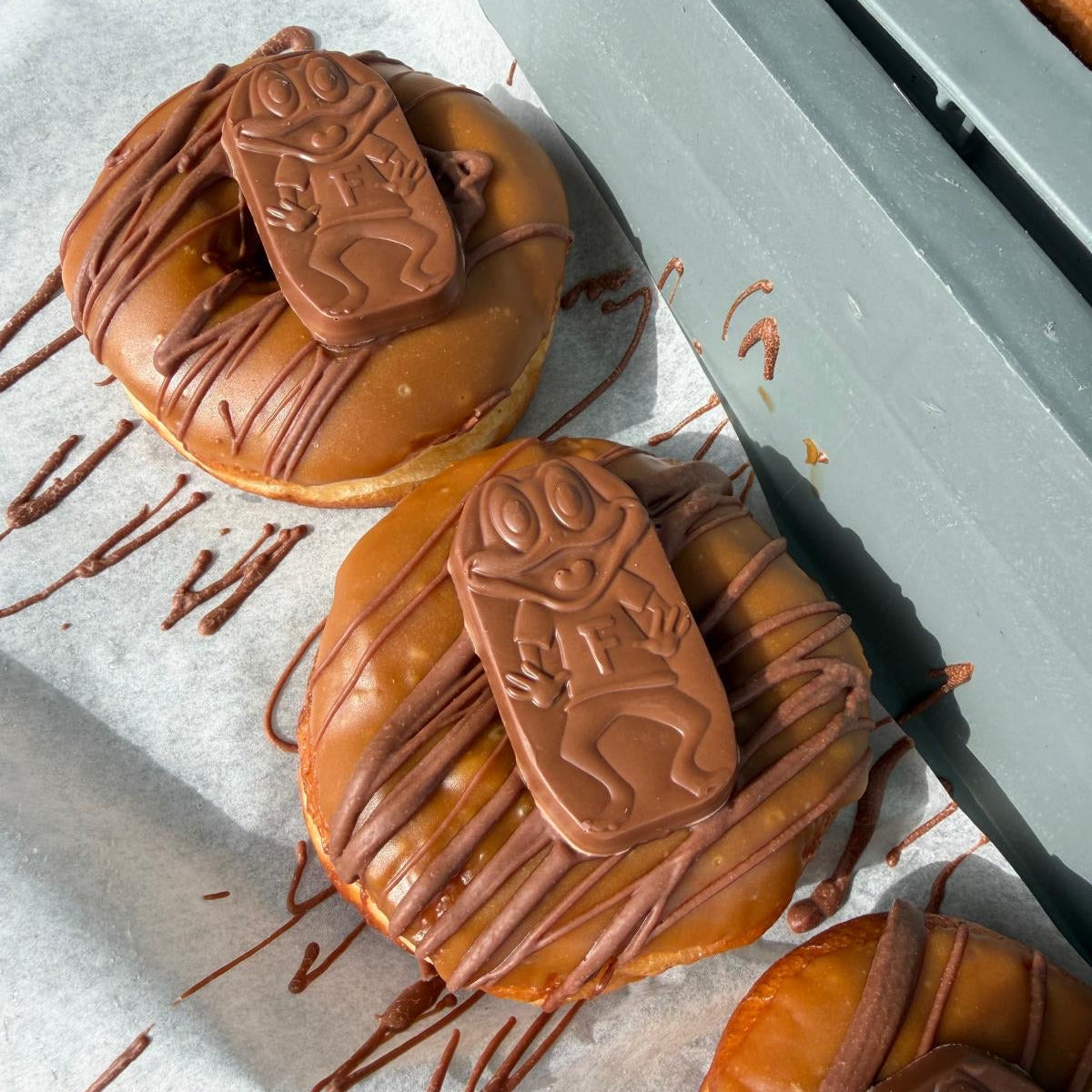 Three glazed donuts topped with chocolate drizzle and a chocolate figure decoration, arranged on parchment paper next to a gray tray.