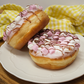Two ring doughnuts topped with white and pink marshmallows with a drizzle of chocolate. In the background is a yellow gingham table cloth.